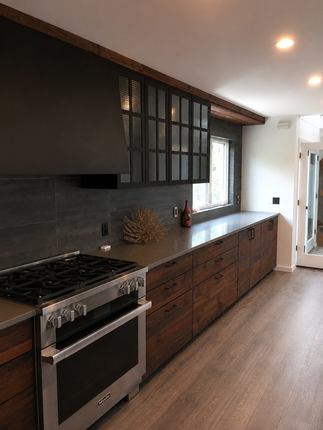Long view of a modern kitchen renovation with reclaimed wood cabinetry, Miele professional range, gray stone tile backsplash, and exposed ceiling beam — New York City by Fuenmayor Solutions LLC